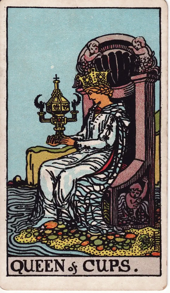 Queen of Cups: A crowned queen sits on a throne carved with cherubs at the edge of the sea, gazing at an ornate covered cup she holds in both hands.