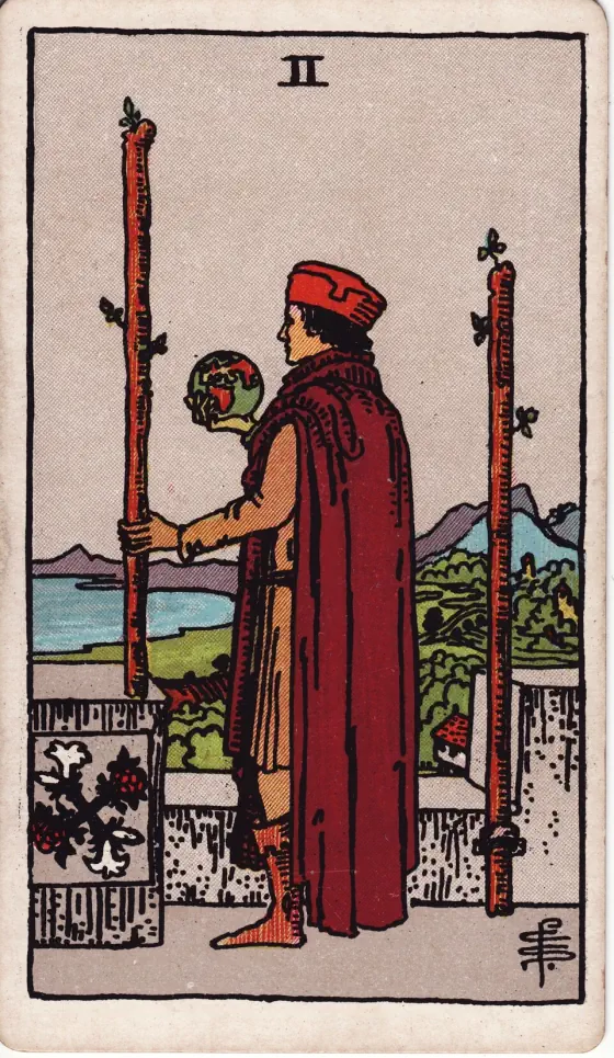 Two of Wands: A tall man looks from a battlemented roof over sea and shore; he holds a globe in his right hand, while a staff in his left rests on the battlement.